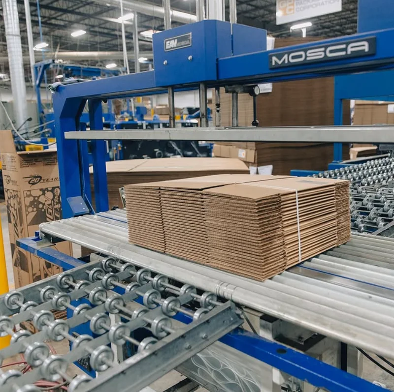 A stack of cardboard boxes on a conveyor belt next to a mosca machine